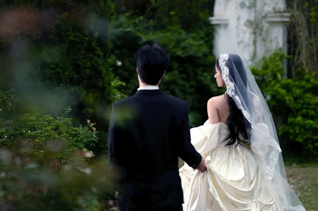 A bride in a white wedding dress and veil walks with a groom in a black suit through a garden.