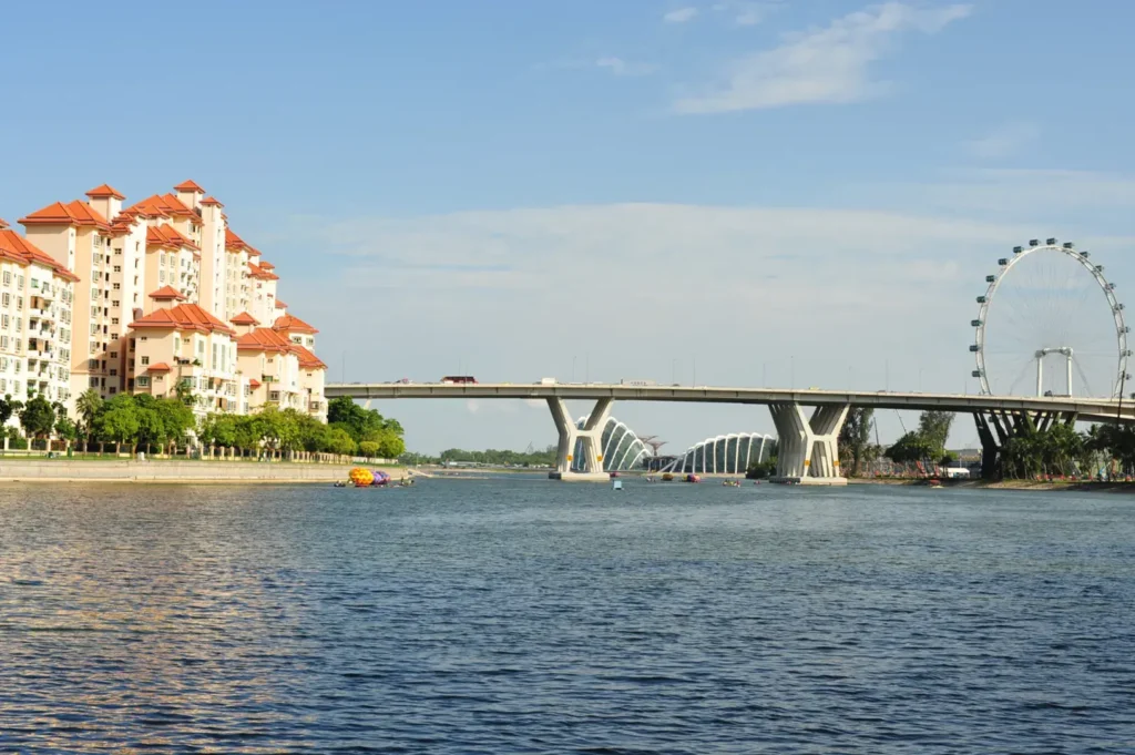 A wide bridge spans a blue body of water, with a cluster of apartment buildings on the left and a Ferris wheel visible in the distance on the right under a clear sky.