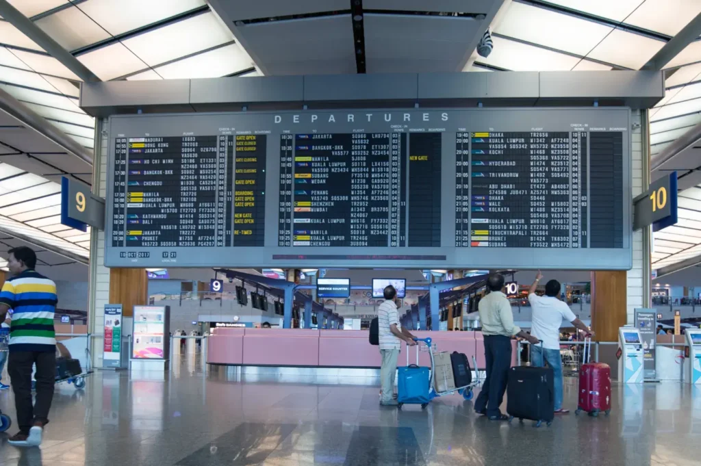 A departures board in an airport with passengers and luggage in the foreground.