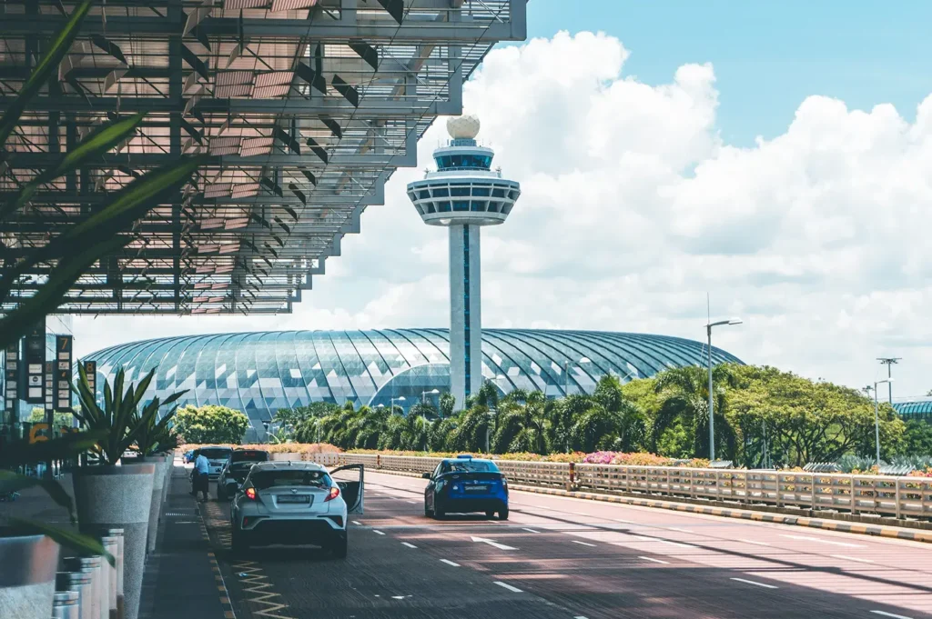 View of the iconic Changi Airport control tower and terminal building in Singapore, with cars driving along the road under a bright, sunny sky.