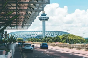 View of the iconic Changi Airport control tower and terminal building in Singapore, with cars driving along the road under a bright, sunny sky.