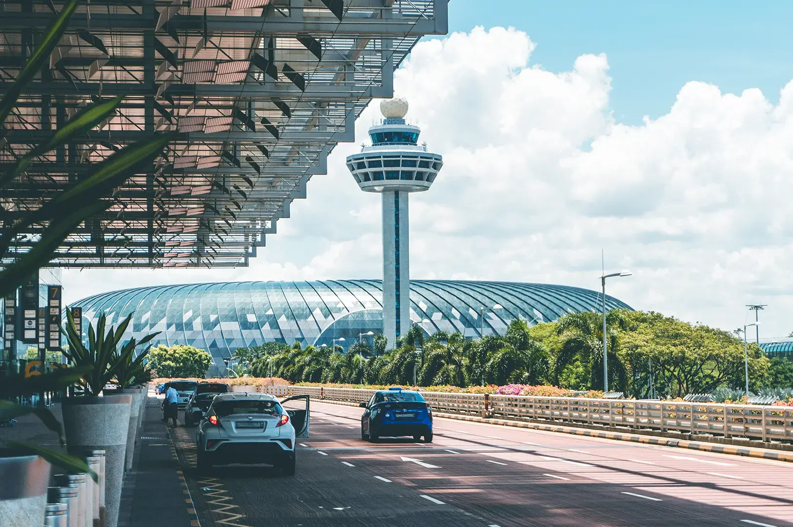 View of the iconic Changi Airport control tower and terminal building in Singapore, with cars driving along the road under a bright, sunny sky.