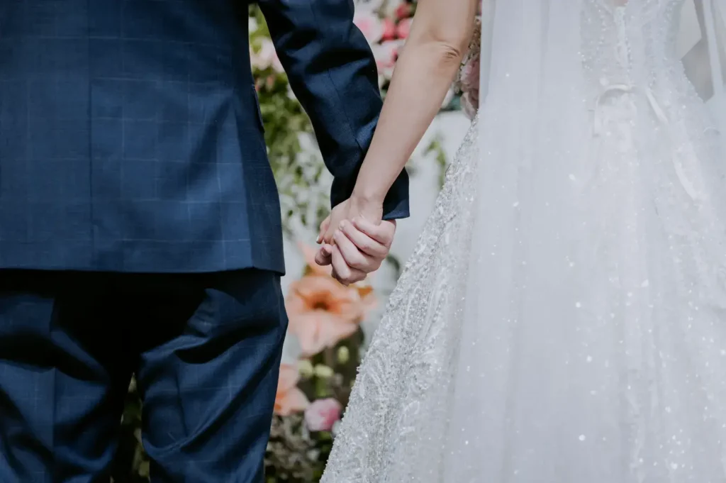 A bride in a sparkly white dress and a groom in a navy blue suit hold hands, with a floral arrangement in the background.