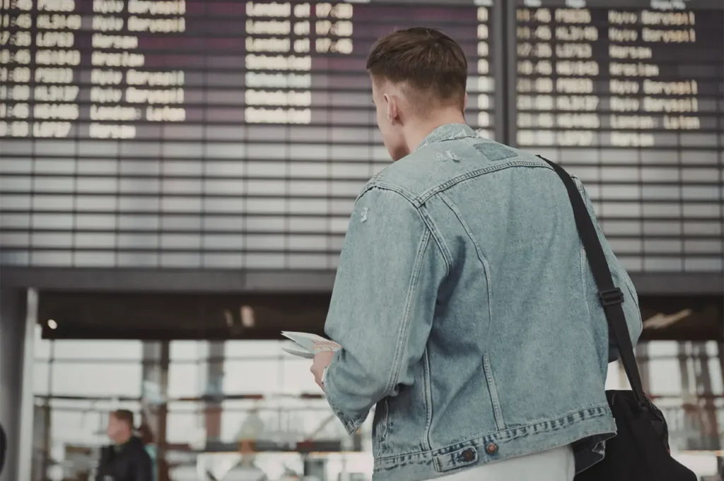 A person in a denim jacket stands in front of a departure board at an airport, holding flight information in their hand.