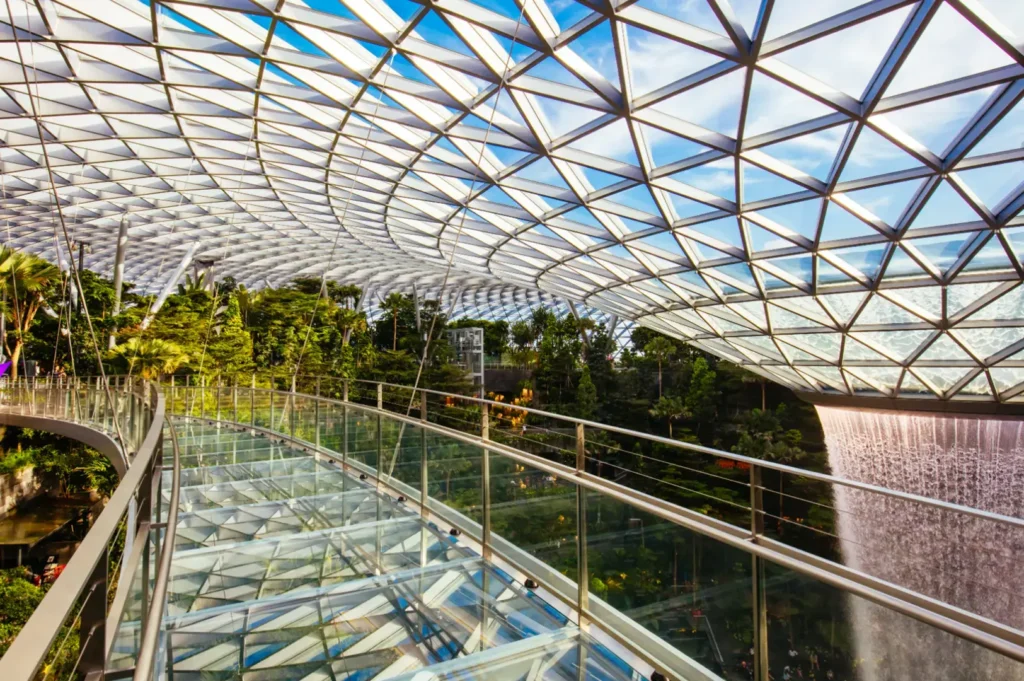 A glass walkway overlooks a lush indoor garden with a waterfall cascading down on the right, all under a geodesic glass roof.