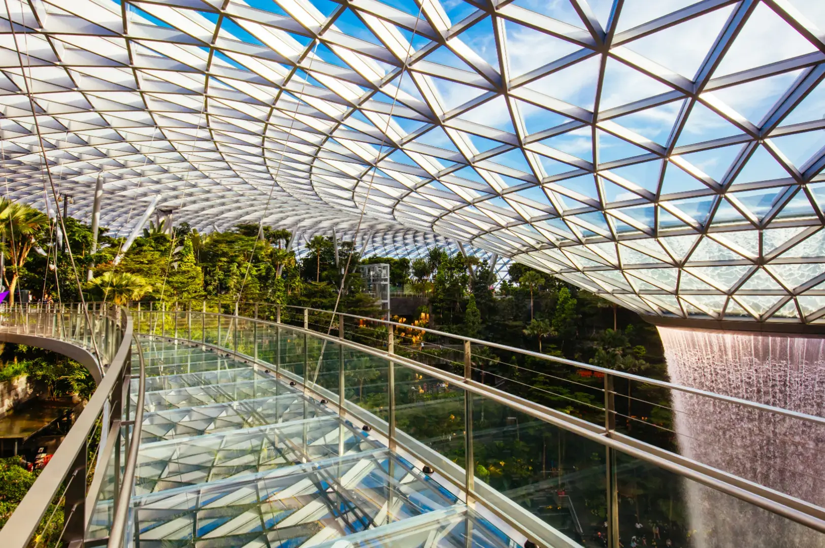 A glass walkway overlooks a lush indoor garden with a waterfall cascading down on the right, all under a geodesic glass roof.