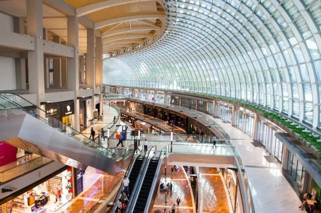 Interior view of a modern shopping mall with multiple levels, escalators, glass railings, and a large arched glass ceiling allowing natural light.