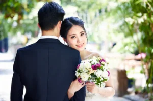 A bride in a white dress smiles at the camera while holding a bouquet and leaning on the shoulder of a groom.