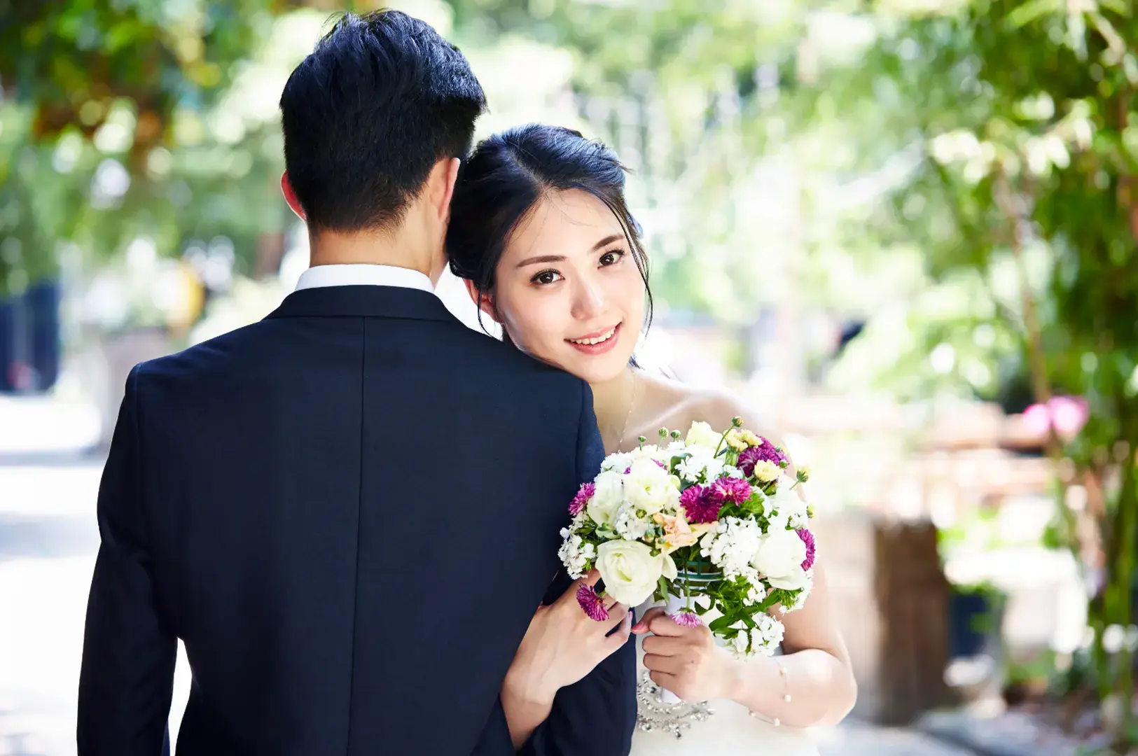 A bride in a white dress smiles at the camera while holding a bouquet and leaning on the shoulder of a groom.