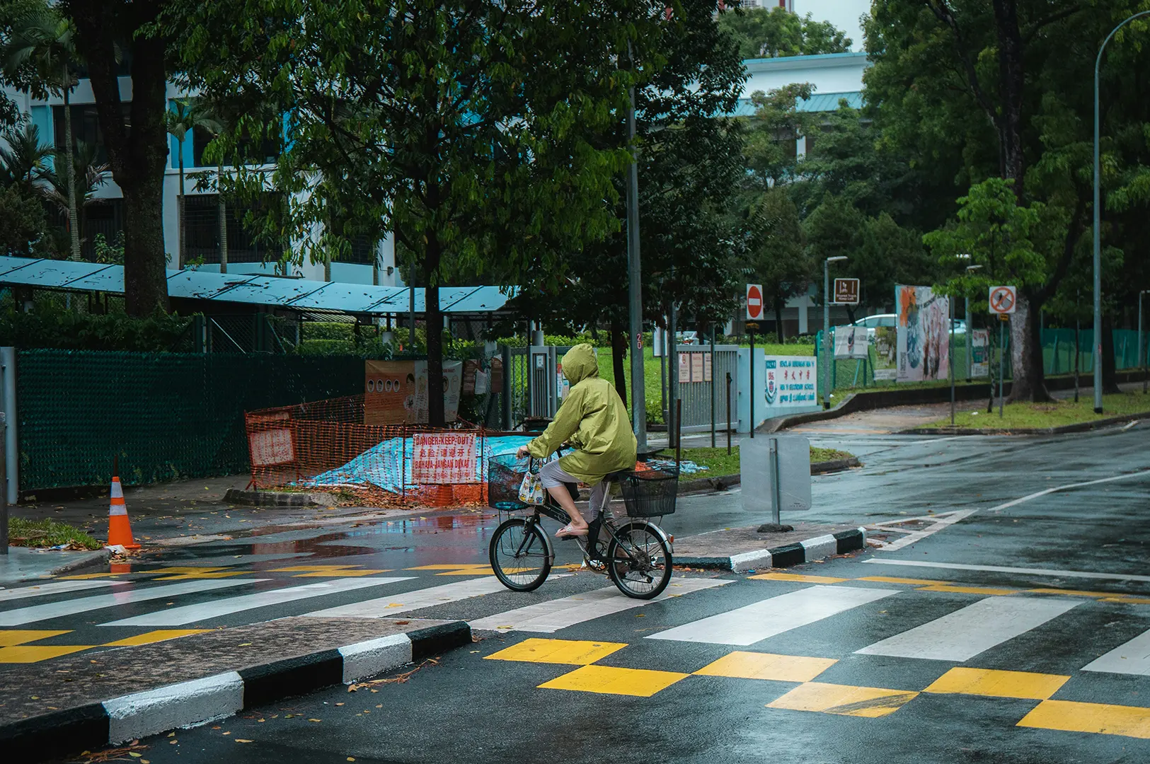 Cyclist in a hooded jacket crossing a wet pedestrian lane on a rainy street.