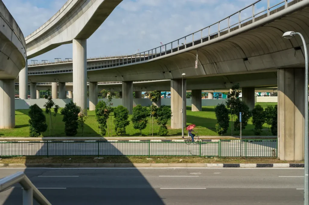 A person on a bicycle rides on a path beneath elevated concrete highway ramps on a sunny day.