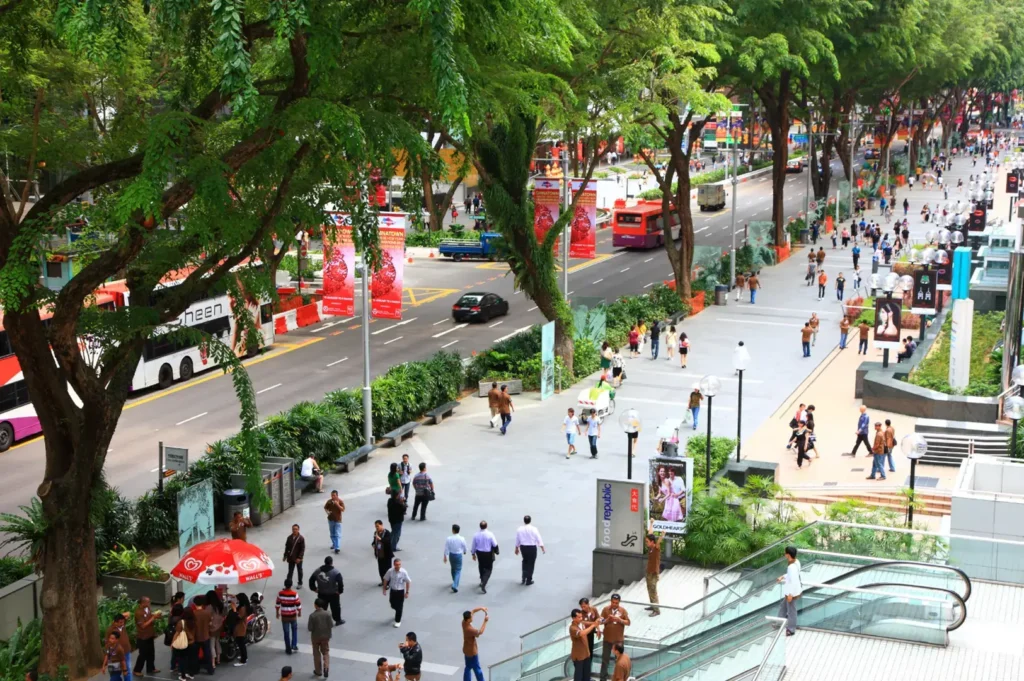 A busy street scene with pedestrians walking on a wide sidewalk lined with trees and shops, while cars and buses travel on the road.