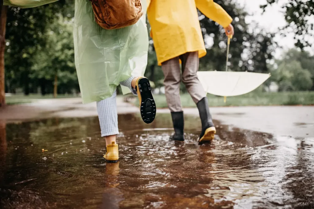 Two people wearing raincoats and boots walk through a large puddle on a wet day. One person carries a closed, light-colored umbrella.