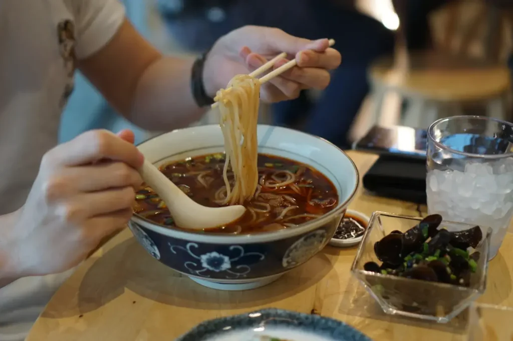 Chopsticks lifting noodles from a bowl of ramen with side dishes.